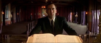 Movie still from “Face/Off” (1997), directed by John Woo – A man sitting in front of an open book; Medium shot, Low angle
