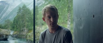 Movie still from “Ex Machina” (2014), directed by Alex Garland – A young blond haired man in front of a window with trees in the background; Medium shot, Over the shoulder angle