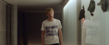 Movie still from “Ex Machina” (2014), directed by Alex Garland – A young man in a white t - shirt standing in a room; Medium shot, Over the shoulder angle