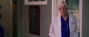 Movie still from “Father Figures” (2017), directed by Lawrence Sher – A man in a white lab coat standing in front of a picture of a dog; Medium shot, Over the shoulder angle