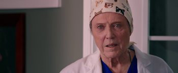 Movie still from “Father Figures” (2017), directed by Lawrence Sher – An older man wearing a white lab coat and a hat with cats on it; Close Up shot, Over the shoulder angle