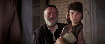 Movie still from “Fiddler on the Roof” (1971), directed by Norman Jewison – An older man wearing a hat and glasses and holding a book; Medium shot, Over the shoulder angle