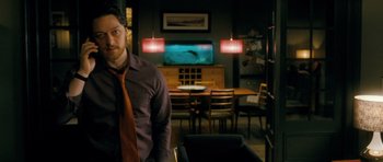 Movie still from “Filth” (2013), directed by Jon S. Baird – A man standing in front of a television in a living room; Medium shot, Over the shoulder angle