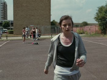 Movie still from “Fish Tank” (2009), directed by Andrea Arnold – A woman is walking in a parking lot with a skateboard; Wide shot, Over the shoulder angle