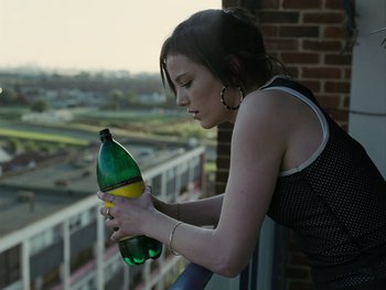 Movie still from “Fish Tank” (2009), directed by Andrea Arnold – A woman is holding a bottle of water on a balcony; Medium shot, High angle