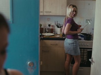 Movie still from “Fish Tank” (2009), directed by Andrea Arnold – A woman standing in a kitchen looking at her cell phone; Medium shot, Over the shoulder angle