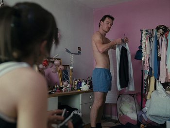 Movie still from “Fish Tank” (2009), directed by Andrea Arnold – A man in blue shorts is holding up a shirt in front of a woman in a pink room; Medium shot, Over the shoulder angle