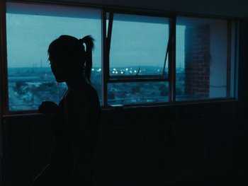Movie still from “Fish Tank” (2009), directed by Andrea Arnold – A woman standing in front of a window at night; Medium shot, Over the shoulder angle