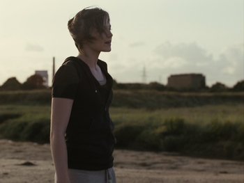 Movie still from “Fish Tank” (2009), directed by Andrea Arnold – A woman standing on a dirt field looking to the side; Medium shot, Over the shoulder angle
