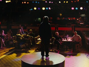 Movie still from “Fish Tank” (2009), directed by Andrea Arnold – A man standing on a stage in front of a group of diners; Wide shot, Over the shoulder angle