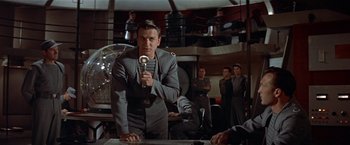 Movie still from “Forbidden Planet” (1956), directed by Fred M. Wilcox – A man holding a microphone while standing next to a table; Medium shot, Low angle