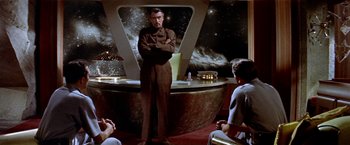 Movie still from “Forbidden Planet” (1956), directed by Fred M. Wilcox – A man standing in front of a group of people in a room; Medium shot, Over the shoulder angle