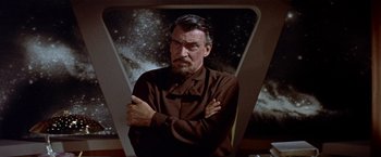 Movie still from “Forbidden Planet” (1956), directed by Fred M. Wilcox – A man with a beard and a brown shirt standing in front of a picture of a galaxy; Medium shot, Low angle