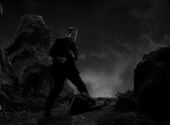 Movie still from “Frankenstein” (1931), directed by James Whale – A black and white photo of a man in a suit and tie standing next to a dead body; Wide shot, Low angle