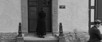 Movie still from “Frantz” (2016), directed by François Ozon – A woman in a long coat and hat standing at the door of a house; Wide shot, High angle