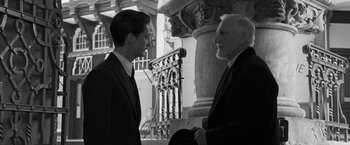 Movie still from “Frantz” (2016), directed by François Ozon – Two men in business suits are standing next to a pillar; Medium shot, Over the shoulder angle