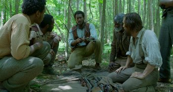Movie still from “Free State of Jones” (2016), directed by Gary Ross – A group of men sitting in the woods holding a stick; Medium shot, Over the shoulder angle