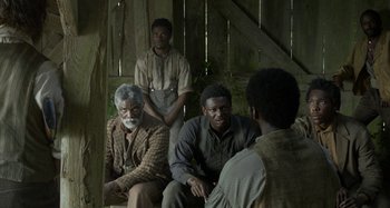 Movie still from “Free State of Jones” (2016), directed by Gary Ross – A group of men sitting on a bench in front of a wooden fence; Medium shot, Over the shoulder angle