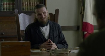 Movie still from “Free State of Jones” (2016), directed by Gary Ross – A man sitting at a table in front of a wooden object; Medium shot, Over the shoulder angle