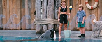 Movie still from “Free Willy” (1993), directed by Simon Wincer – A woman in a black top and black shorts holding an object in front of an orca; Wide shot, Over the shoulder angle