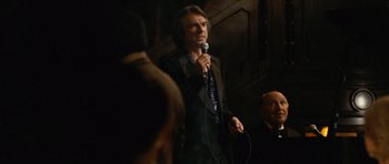 Movie still from “Frost/Nixon” (2008), directed by Ron Howard – A man holding a microphone while standing in front of a group of people; Medium shot, Over the shoulder angle