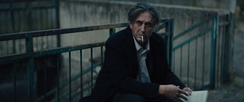 Movie still from “Hellraiser” (2022), directed by David Bruckner – An older man sitting on a railing with a cigarette in his mouth; Medium shot, Low angle