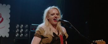 Movie still from “Her Smell” (2018), directed by Alex Ross Perry – A woman with long blonde hair singing into a microphone; Medium shot, Low angle