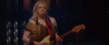 Movie still from “Her Smell” (2018), directed by Alex Ross Perry – A woman holding a guitar while standing in front of a microphone; Medium shot, Over the shoulder angle