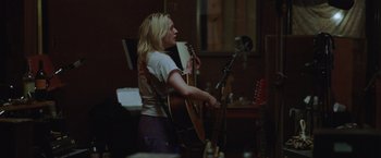 Movie still from “Her Smell” (2018), directed by Alex Ross Perry – A woman holding a guitar in front of a microphone in a room; Medium shot, Over the shoulder angle