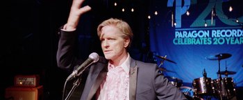 Movie still from “Her Smell” (2018), directed by Alex Ross Perry – A man in a suit and tie holding a microphone; Medium shot, Low angle