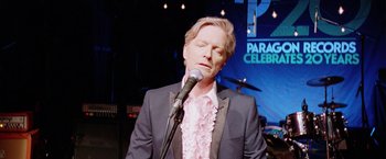 Movie still from “Her Smell” (2018), directed by Alex Ross Perry – A man in a suit and pink shirt is singing into a microphone; Close Up shot, Over the shoulder angle