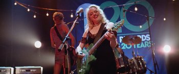 Movie still from “Her Smell” (2018), directed by Alex Ross Perry – A woman in a black dress playing an electric guitar; Medium shot, Low angle