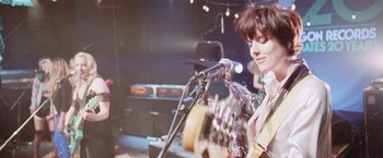 Movie still from “Her Smell” (2018), directed by Alex Ross Perry – A person playing a guitar on a stage; Medium shot, Over the shoulder angle