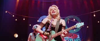 Movie still from “Her Smell” (2018), directed by Alex Ross Perry – A woman in a black dress is playing a guitar; Medium shot, Low angle