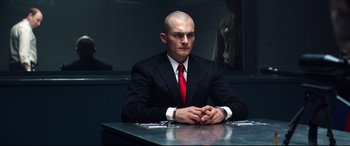 Movie still from “Hitman: Agent 47” (2015), directed by Aleksander Bach – A man sitting at a table in a suit and red tie; Medium shot, Low angle