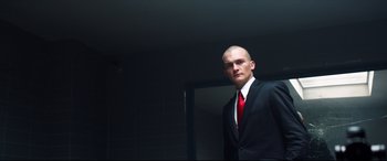 Movie still from “Hitman: Agent 47” (2015), directed by Aleksander Bach – A man in a suit and red tie standing next to a mirror; Medium shot, Low angle