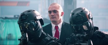 Movie still from “Hitman: Agent 47” (2015), directed by Aleksander Bach – A man in a suit and tie standing next to two other men in helmets; Close Up shot, Low angle