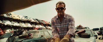 Movie still from “Get the Gringo” (2012), directed by Adrian Grunberg – A man in a plaid shirt and gloves is sitting in front of a car; Medium shot, Low angle