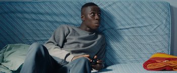Movie still from “Girlhood” (2014), directed by Céline Sciamma – A man sitting on a couch holding a remote control; Medium shot, High angle