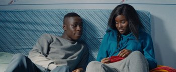 Movie still from “Girlhood” (2014), directed by Céline Sciamma – A man and a woman sitting next to each other on a couch; Medium shot, Over the shoulder angle