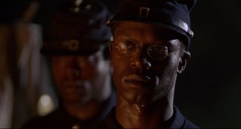 Movie still from “Glory” (1989), directed by Edward Zwick – A man wearing a hat and glasses is looking at the camera; Close Up shot, Over the shoulder angle