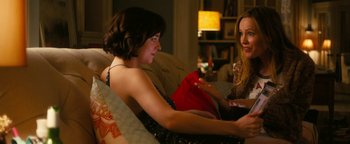 Two women are sitting on a couch and one of the women is holding a red bag