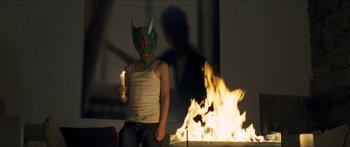 Movie still from “Goodnight Mommy” (2014), directed by Severin Fiala – A person wearing a mask and holding a lit candle in front of a fire; Medium shot, Over the shoulder angle