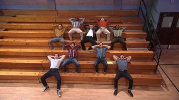 Movie still from “Grease Live!” (2016), directed by Thomas Kail – A group of men sitting on top of a wooden bleacher; Wide shot, High angle