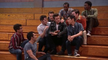 Movie still from “Grease Live!” (2016), directed by Thomas Kail – A group of young men sitting on top of bleachers looking at a cell phone; Wide shot, High angle