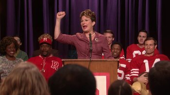 Movie still from “Grease Live!” (2016), directed by Thomas Kail – A woman is giving a speech in front of a group of people; Medium shot, Low angle