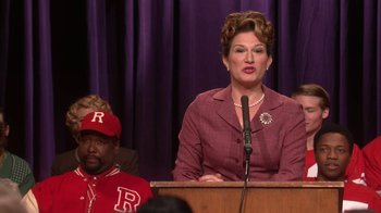 Movie still from “Grease Live!” (2016), directed by Thomas Kail – A woman is speaking at a podium in front of a crowd; Medium shot, Over the shoulder angle