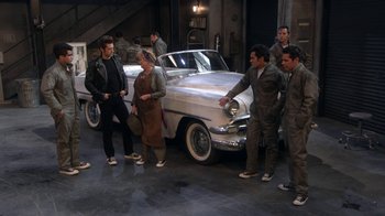 Movie still from “Grease Live!” (2016), directed by Thomas Kail – A group of men standing around a silver car; Wide shot, High angle