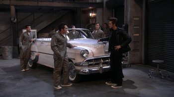 Movie still from “Grease Live!” (2016), directed by Thomas Kail – A group of men standing next to an old car; Wide shot, High angle