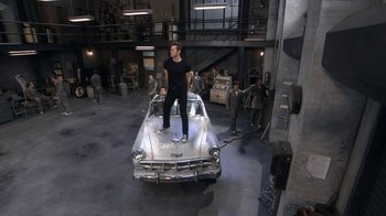 Movie still from “Grease Live!” (2016), directed by Thomas Kail – A man standing on the hood of an old car; Wide shot, High angle